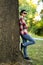 Attractive teenage boy in checkered shirt, blue jeans, sunglasses and baseball cap lean on a tree waiting for someone or