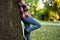 Attractive teenage boy in checkered shirt, blue jeans, sunglasses and baseball cap lean on a tree waiting for someone or