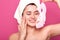 Attractive tender young lady standing  over pink background in studio, holding face cosmetic mask in one hand, touching