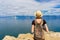 Attractive tween tourist girl in hat and backpack standing on cliff top and admiring beautiful landscape of blue sky and Baikal