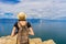 Attractive tween tourist girl in hat and backpack standing on cliff top and admiring beautiful landscape of blue sky and Baikal
