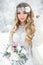 Attractive winter girl in a white outfit in a snow forest with beautiful flowers on her head and a bouquet