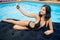 Attractive woman in bikini smiling and taking selfie photo on the phone on a mattress in the pool at the resort