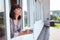 Attractive woman holding a cup of tea while staying in window frame of her country house on second floor, side view