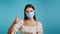 Attractive woman in protective respirator showing thumb up sign over blue background. Positive young girl in mask smiles