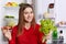 Attractive woman wears fashionable red blouse, stands near opened fridge, holds lettuce and sandwhich, going to prepare delicious