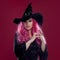 Attractive woman in witches hat and costume with red hair performs magic on pink background. Halloween, horror theme.