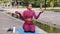 Attractive young adult woman fitness instructor sitting on yoga mat in summer park holds apple and cake, cupcake, trying