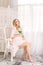 Attractive young blonde pregnant woman sitting on chair in white nightgown. Happy glamour girl at home.