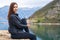 Attractive young brunette woman sits on stone pier and gazes thoughtfully out to sea, beautiful mountain range on