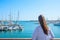 Attractive Young Caucasian Woman with Long Hair Boho Tunic Stands at Beach Looks at Yachts Sailing Boats Moored in Marina. Sea Sky