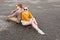 An attractive young couple in sunglasses sit back to back on their longboard in a suburban parking lot against a gray