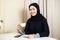 Attractive young female muslim holding coffee mug cup sitting at home and smiling to the camera