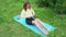 Attractive young fitness woman using rubber band exercising her back in garden on grass. Smiling girl in sportswear does