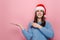 Attractive young girl keeps palm raised, points with forefinger against pink wall for advertisement, dressed in Christmas red hat