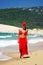 Attractive young girl in red sarong, bikini and baseball cap walking along crowded white sandy beach