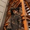 Attractive young hipster man in knitted hat in camouflage shirt in sunglasses is posing standing near the iron orange stairs