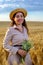 Attractive young long-haired woman in straw hat smiling and holds a wild flower bouquet in wheat field at sunrise