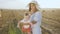 Attractive young mother in big hat and lightweight dress playing with baby boy on the field with bales. Mom with little