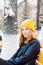 Attractive young woman with blue eyes and blond hair in a yellow knitting hat sitting on a yellow bench in the winter park