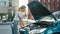 Attractive young woman examining, looking at her broken car with open hood while standing on the city street