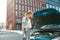 Attractive young woman examining, looking at her broken car with open hood while standing on the city street