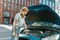 Attractive young woman examining, looking at her broken car with open hood while standing on the city street