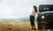 Attractive Young Woman Traveler Enjoying The Sea View, Leaning Back On a Classic Car SUV. Adventure Travel Concept and Lifestyle