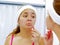 Attractive young woman wearing pink top and white headband, touching face with skincare tool, looking in mirror