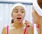 Attractive young woman wearing pink top and white headband, touching face with skincare tool, looking in mirror