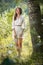 Attractive young woman in white short dress posing near a tree in a sunny summer day. Beautiful girl enjoying the nature