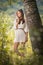 Attractive young woman in white short dress posing near a tree in a sunny summer day. Beautiful girl enjoying the nature