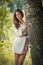 Attractive young woman in white short dress posing near a tree in a sunny summer day. Beautiful girl enjoying the nature