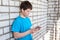 Attractive youngster guy wearing blue t-shirt holding smartphone in hands, browsing social media, leaning brick wall