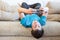 Attractive youngster guy wearing blue t-shirt lying on his back on couch and holding smartphone in hands, browsing social media