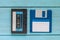 An audio cassette in a black plastic case and a blue floppy disk lie on a blue wooden background