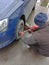 The auto mechanic sits down next to the wheel of the car and unscrews the bolts on it with a special key to replace it