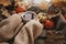 Autumn hygge. Hands in cozy sweater holding warm cup of tea with stylish pumpkins, fall leaves, lights on brown scarf on