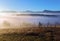 Autumn landscape of high mountains, orange coloured trees, fog. Sun rays enlighten the lawn with dry grass. Blue sky.