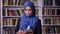 Awesome arabian woman in dark blue hijab is holding book in library, standing calmly and confident glance at camera