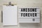 `Awesome forever` word on modern board, clipboard with sheet of paper on a white wooden surface, top view. From above, flat lay,