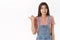 Awkward and slightly worried cute brunette female in overalls, t-shirt, pointing thumb left, clench teeth and smiling