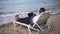 Azure blue foamy Mediterranean sea waves splashing on sandy beach with blurred African American man lying on sunbed