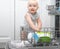 Baby blonde boy with blu eyes helping in the kitchen using dishwasher. white kitchen interior.
