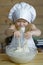 Baby or child in cook uniform with chef hat and apron kneading dough with flour in glass bowl in kitchen.