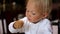 Baby eating food in restaurant. Blonde blue-eyed child in highchair taking piece of food by hand from a plate putting it
