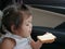 A baby girl, 2 years old, learning to eat, by herself, a sheet of white bread with sweetened condensed milk on top in a car during