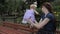 A baby girl climbing on the bench back with her legs, a girl holding her by the buttocks and arm pit.