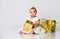 Baby girl in dress and headband, barefoot. She holding golden gift box, sitting on floor isolated on white. Close up, copy space