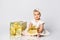 Baby girl in dress and headband, barefoot. She holding golden gift box, sitting on floor isolated on white. Close up, copy space